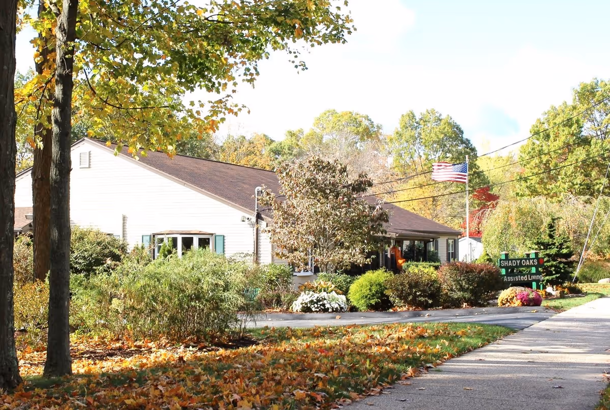 Exterior view of Shady Oaks Assisted Living LLC building surrounded by trees with autumn foliage, bushes, and a walkway leading to the entrance. An American flag is flying on a flagpole near the building.