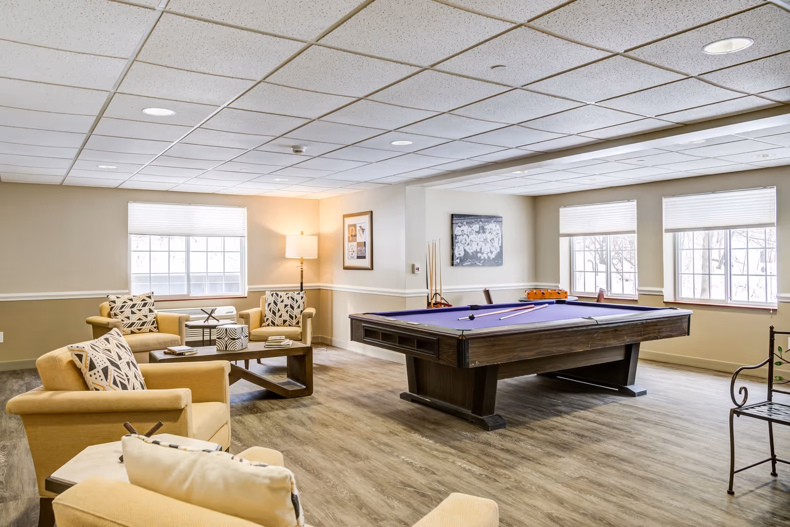 A bright and spacious senior living common area featuring a pool table with purple felt, several beige armchairs with patterned cushions, a wooden coffee table with books and decorative items, large windows with white blinds, and framed artwork on the walls.