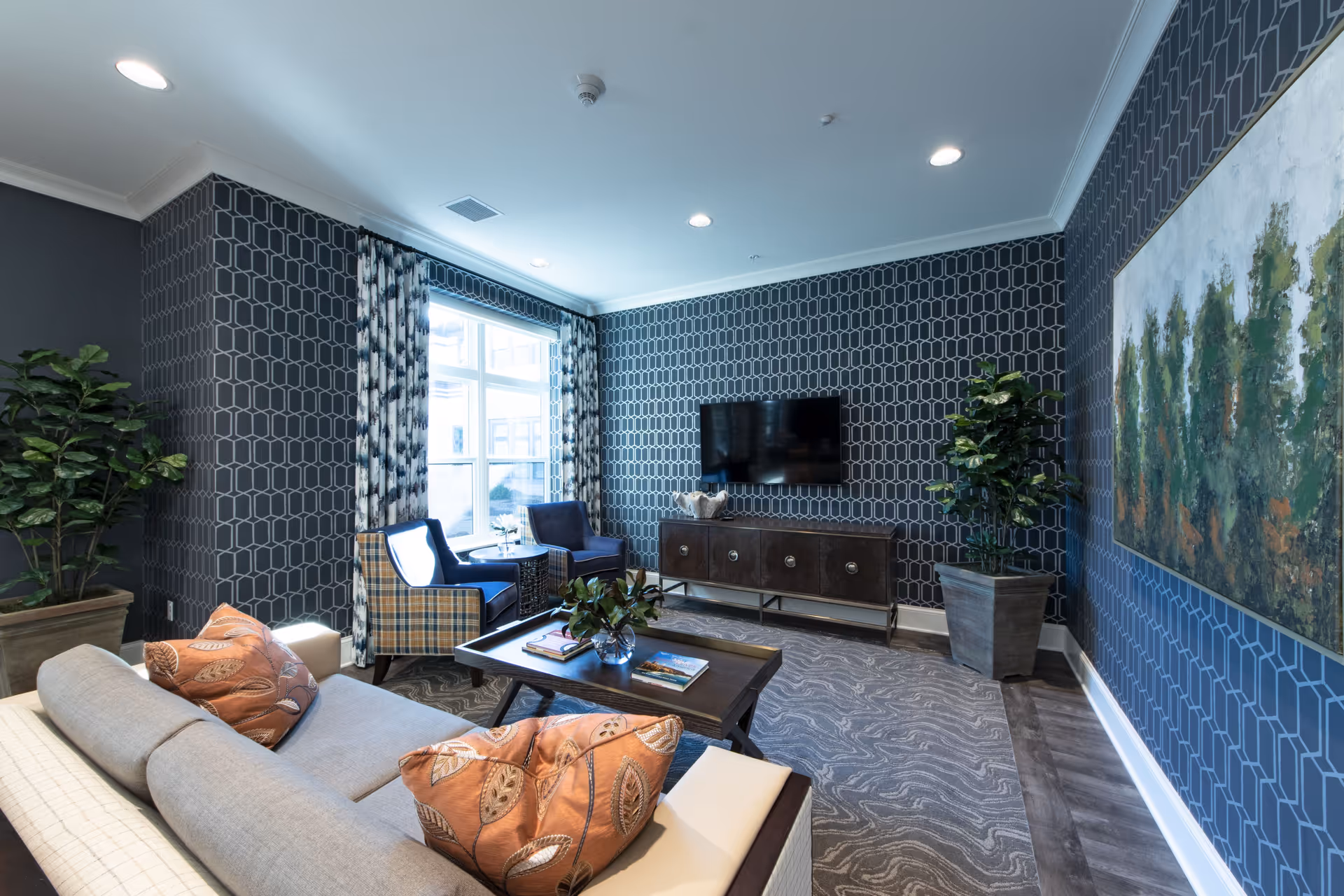 A modern living room with patterned dark blue wallpaper, a large window with patterned curtains, a wall-mounted flat-screen TV above a dark wooden console, two blue and plaid armchairs, a beige sofa with orange patterned pillows, a wooden coffee table with books and a small plant, two large potted plants, and a large painting of trees on the wall.