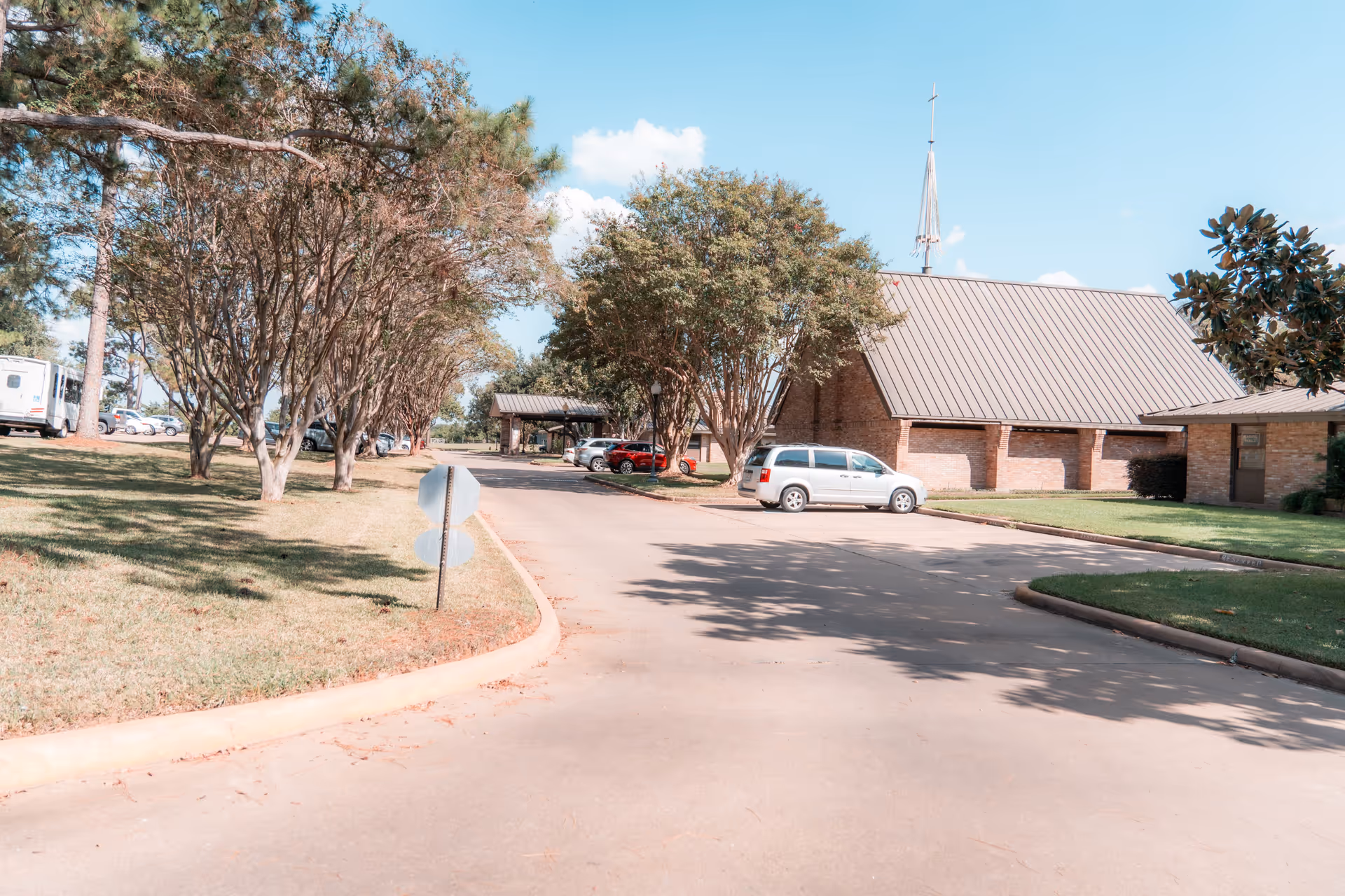 A paved driveway lined with trees and grass leads to a brick building with a steep metal roof and a tall spire. Several cars are parked along the driveway under a clear blue sky.