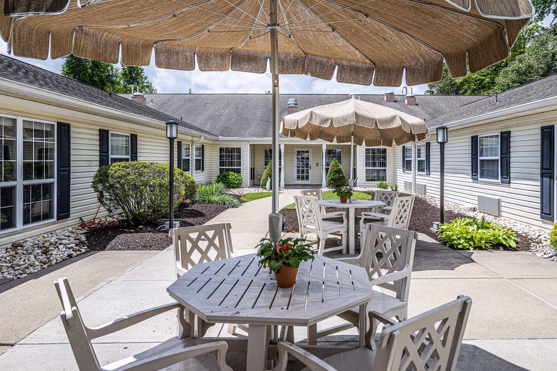 Outdoor courtyard area with beige patio umbrellas shading white wooden tables and chairs. The courtyard is surrounded by a single-story building with white siding and black shutters. There are small landscaped garden beds with bushes and plants along the building walls.