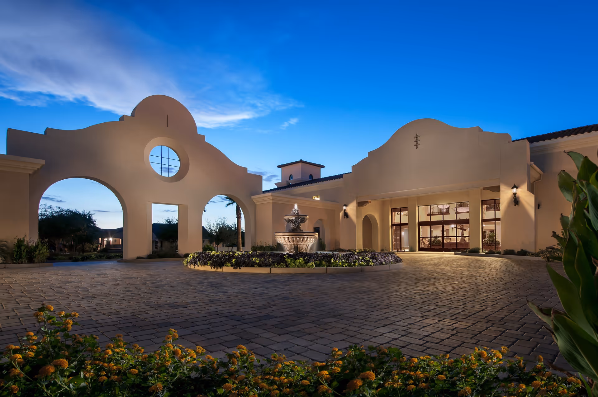 Exterior view of Clearwater Agritopia facility at dusk, featuring a large arched entrance, a central water fountain surrounded by landscaping, and warm lighting illuminating the building's facade.