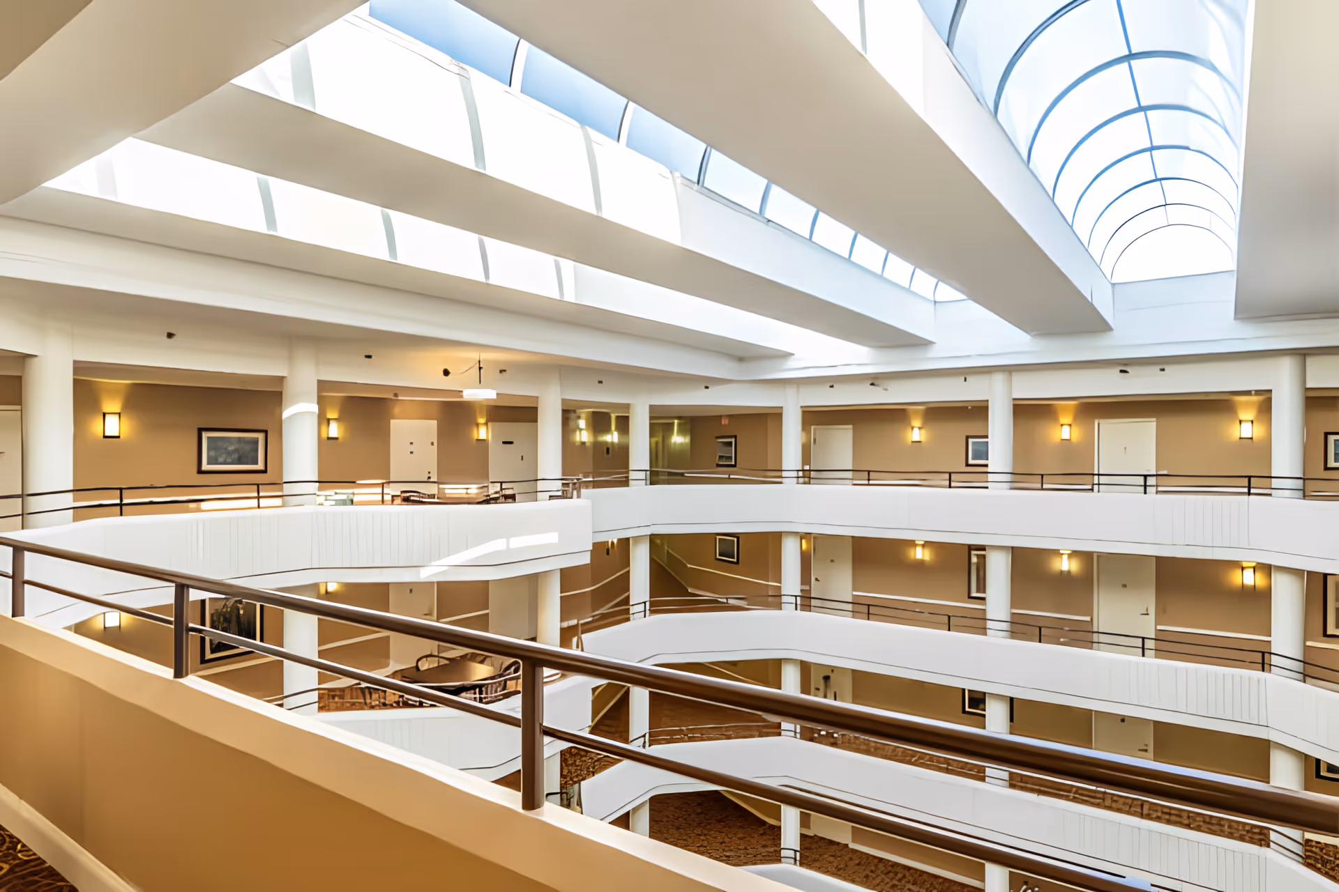 Sunlit multi-level interior atrium with curved balconies, metal railings, seating areas, and a large arched skylight.