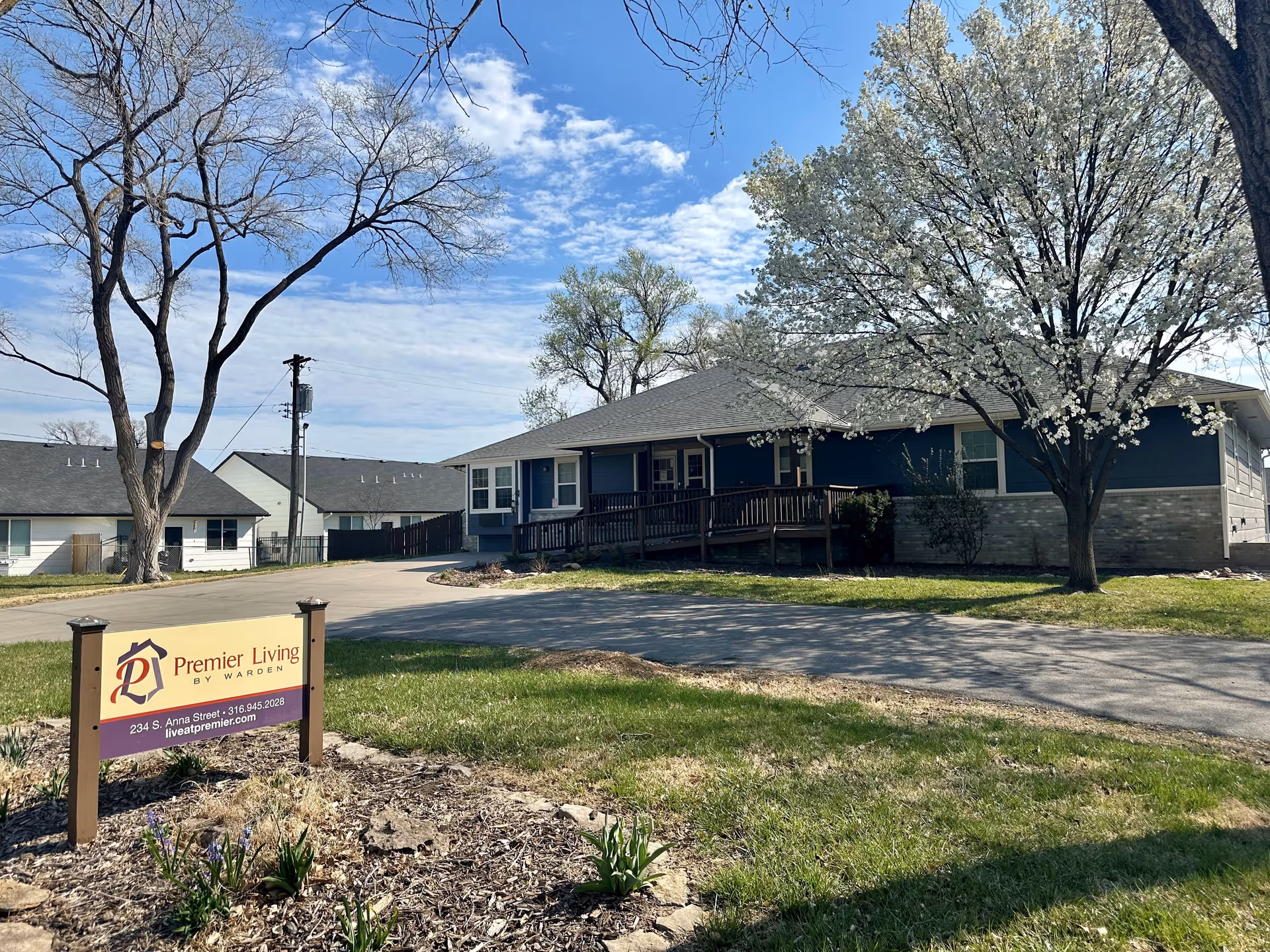Exterior view of Premier Living by Warden, LLC facility showing a single-story building with a ramp entrance, surrounded by trees including one with white blossoms, a driveway, and a sign with the facility's name and contact information in the foreground.
