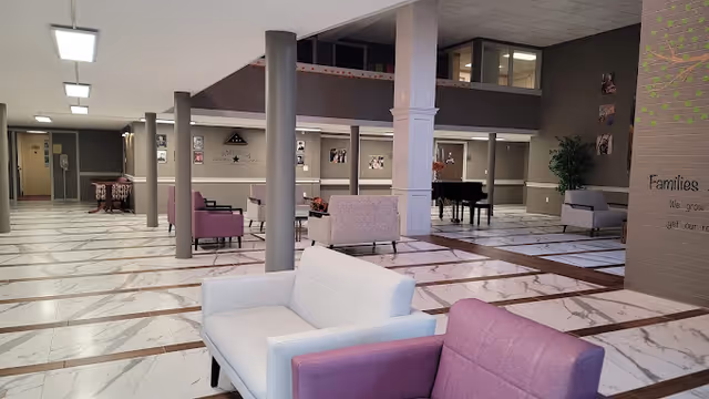 Spacious interior common area with white and purple armchairs arranged on a white marble floor with brown accents. The room features multiple columns, a grand piano in the background, and framed pictures on the walls. There is a partial view of a wall with text and a potted plant on the right side.