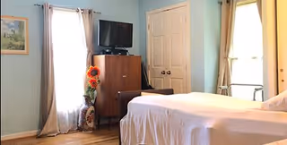 A bedroom with a bed covered in white sheets, a wooden cabinet with a flat-screen TV on top, a window with beige curtains, a vase with sunflowers, and a closed white door.