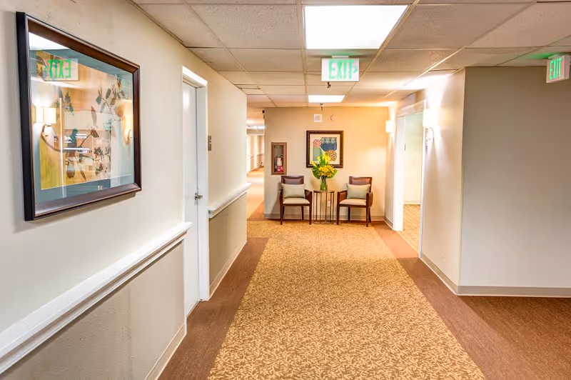 A well-lit hallway in a senior living facility with beige walls and patterned carpet. Two wooden chairs with cushions and a small table with a flower arrangement are placed against the far wall beneath a framed artwork. Exit signs are visible on the ceiling and walls, and a fire extinguisher is mounted on the wall near the chairs.