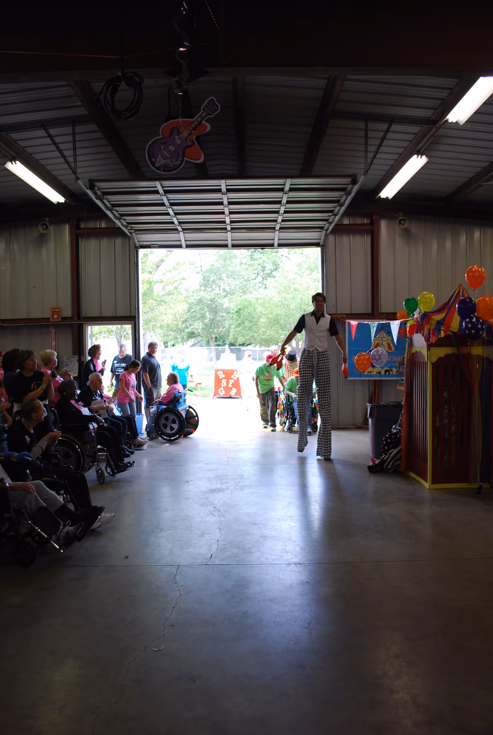 A group of people, some in wheelchairs, seated and standing inside a large garage-like space with an open door leading outside. A performer on stilts is walking towards the camera, and colorful balloons and a small carnival-style booth are visible on the right side. Outside, trees and a bright sky can be seen.