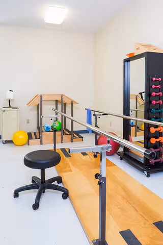 A small physical therapy or exercise room with parallel bars on a wooden platform, a black rolling stool, colorful exercise balls, and a rack holding various small dumbbells next to a full-length mirror.