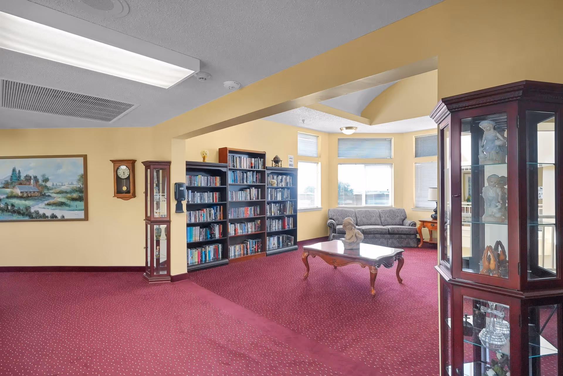 A cozy common area with yellow walls and red carpet featuring a wooden coffee table with a decorative bust, a gray upholstered sofa near large windows, bookshelves filled with books, a wall clock, a painting of a countryside scene, and a glass display cabinet with various figurines.