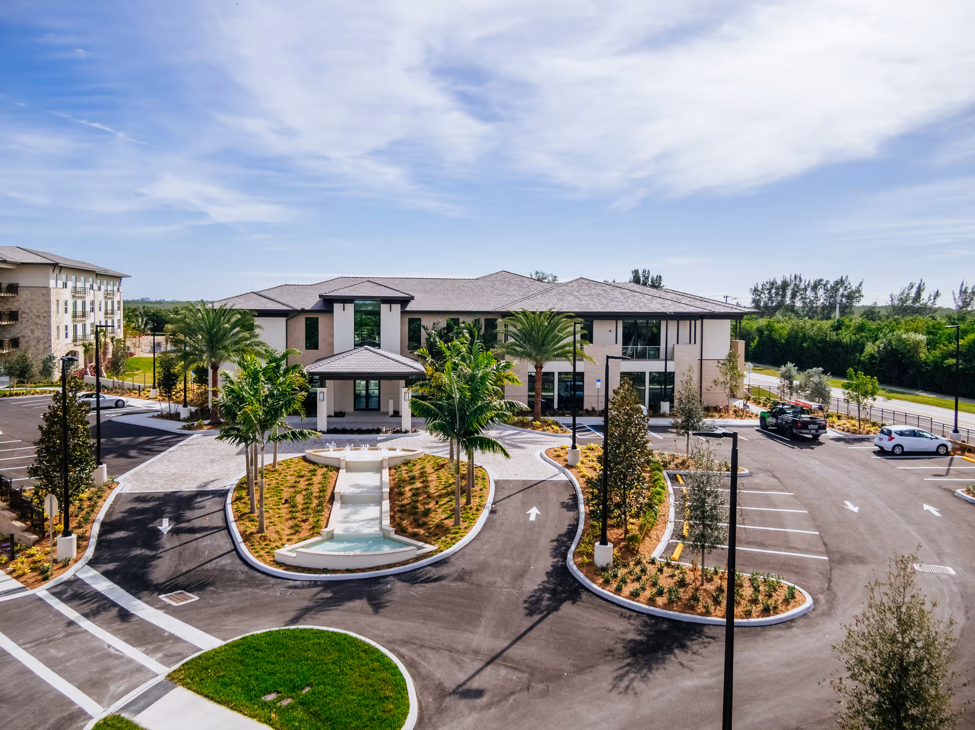 Exterior view of the Shell Point Welcome Center building with a driveway, landscaped garden featuring palm trees and a fountain, and a parking lot with a few cars under a partly cloudy sky.