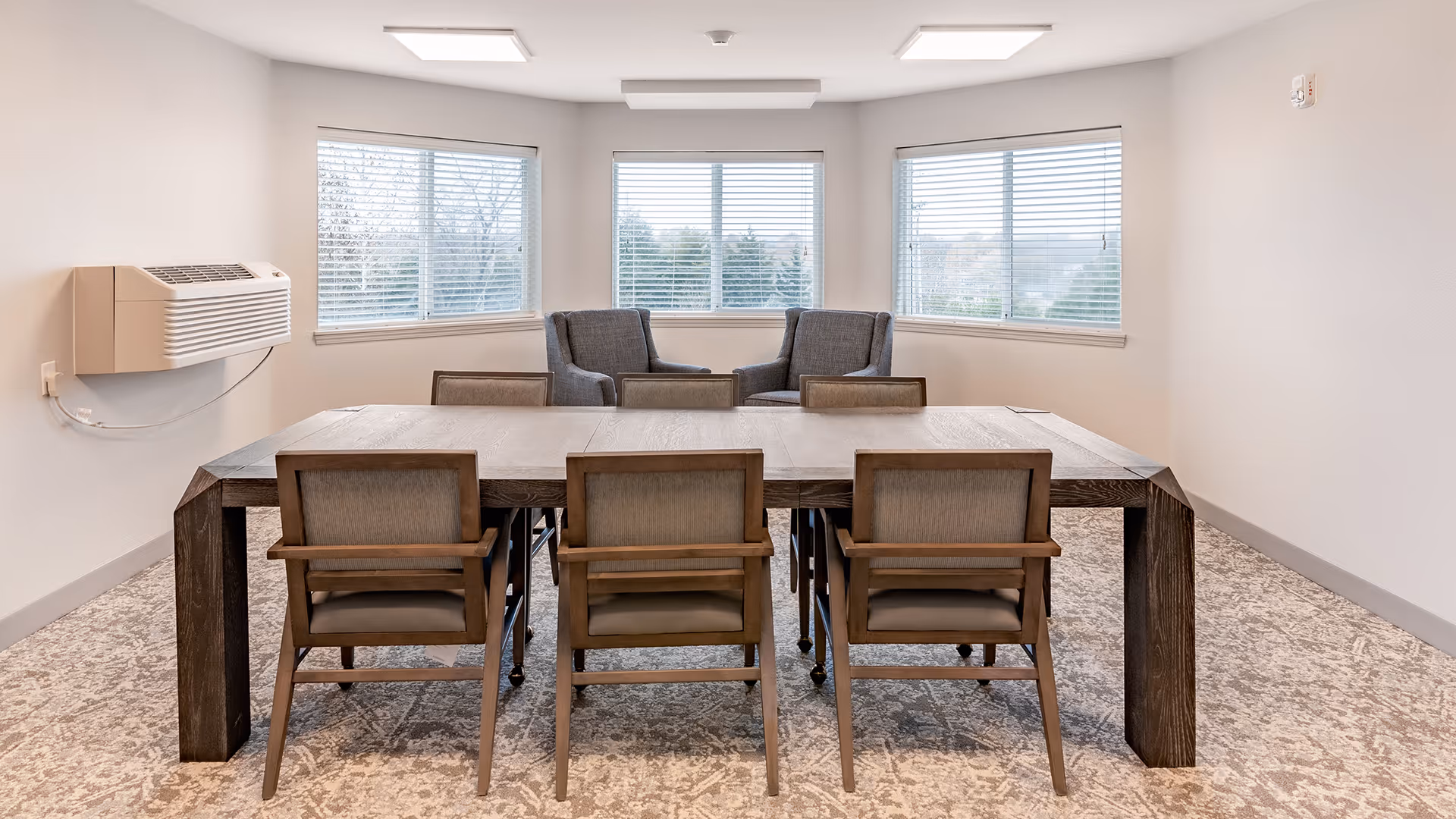 A bright room with a large wooden table surrounded by six cushioned chairs. In the background, there are three large windows with blinds partially open, and two gray armchairs positioned near the windows. The room has light-colored walls and carpeted flooring, with a wall-mounted air conditioning unit on the left side.