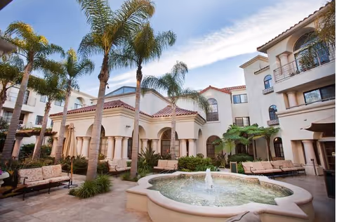 Outdoor courtyard area of a senior living facility with palm trees, cushioned benches, umbrellas, and a central water fountain surrounded by a beige stone patio. The building has a Mediterranean architectural style with arched windows and balconies.