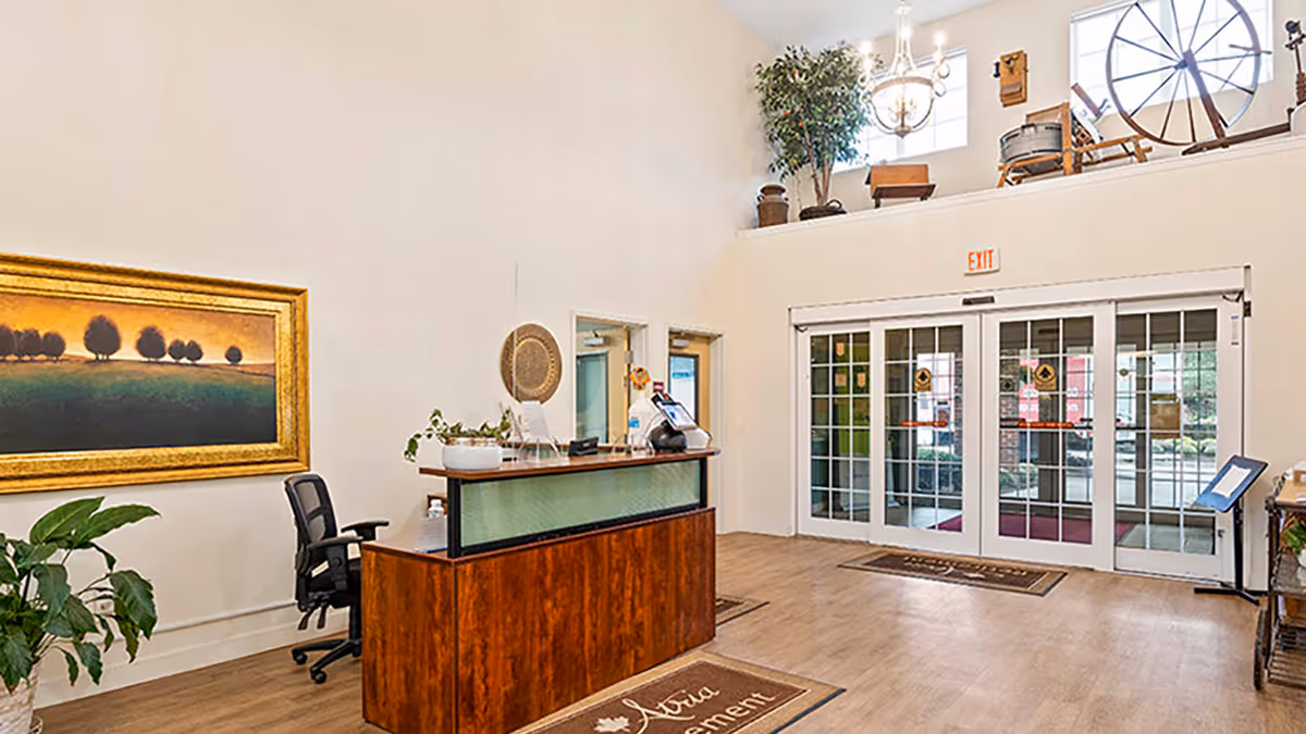 Reception area of a senior living facility with a wooden front desk, an office chair, a large framed painting of trees on the wall, potted plants, and glass double doors leading outside. Above the doors is a high shelf displaying antique items including a spinning wheel and a wooden chair, with large windows letting in natural light.
