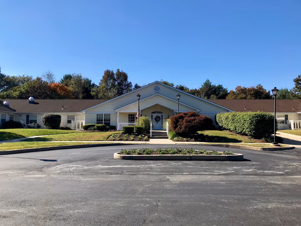 Front exterior view of a single-story building with a peaked roof and white siding, surrounded by neatly trimmed bushes and trees under a clear blue sky. There is a circular driveway with a small landscaped island in the center and two lampposts near the entrance.