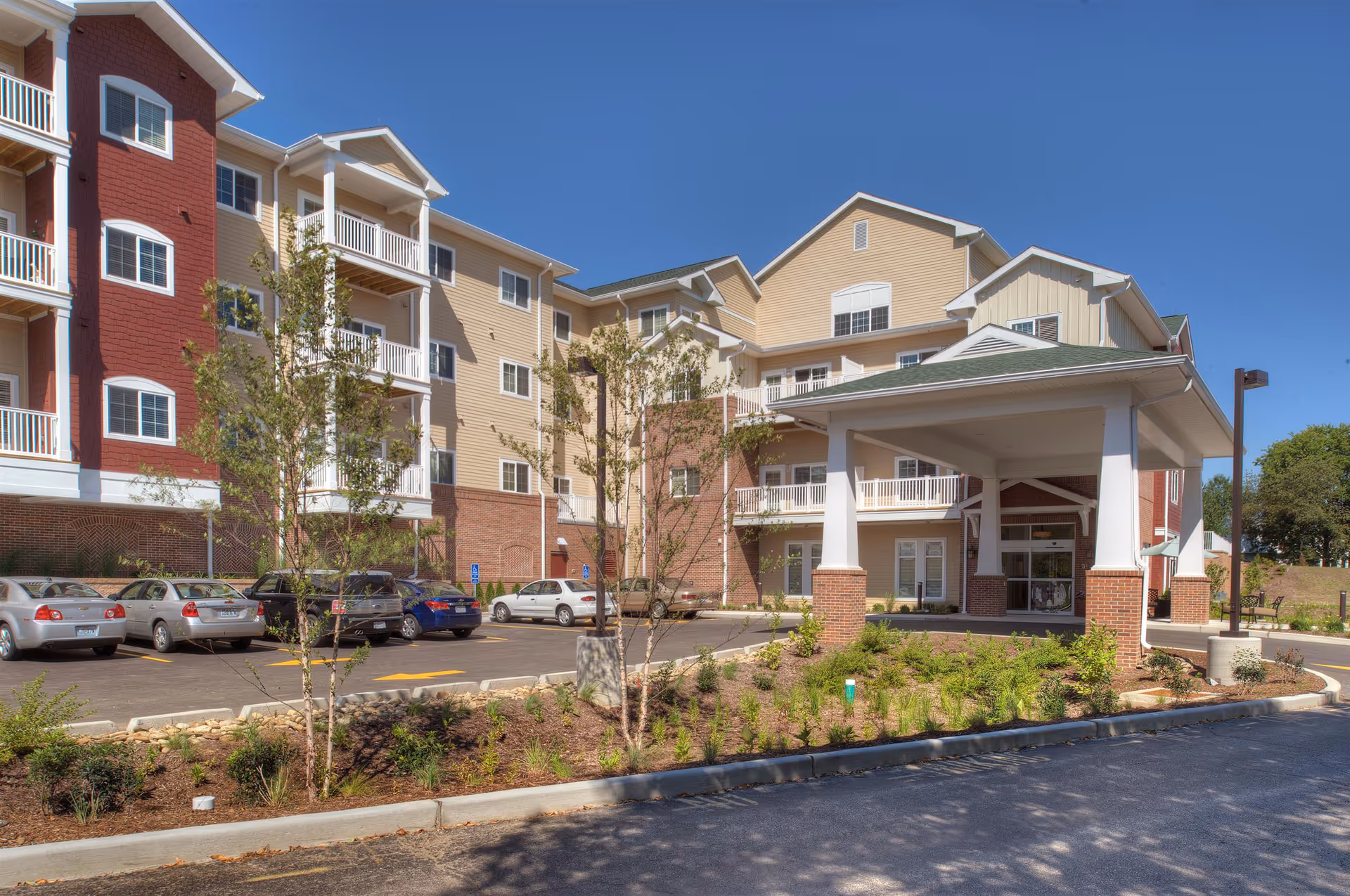 Exterior view of Nazareth Living Center, a multi-story residential building with beige and red siding, white balconies, and a covered entrance. Several cars are parked in the parking lot in front of the building, and there are small trees and landscaped areas around the entrance.