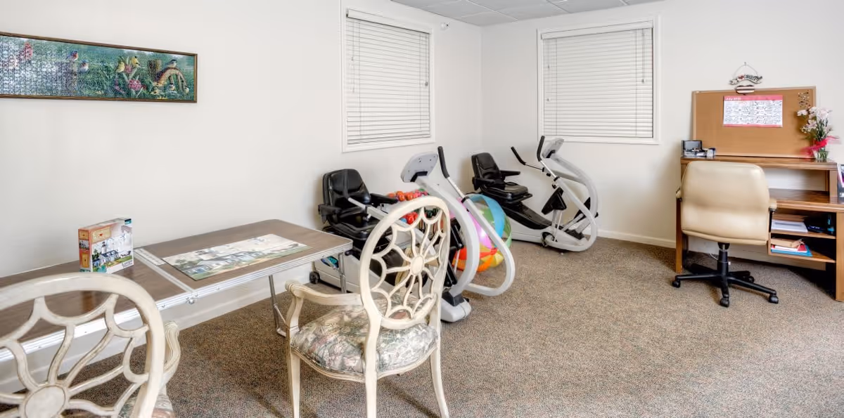 A small room with exercise equipment including two recumbent bikes and colorful exercise balls. There is a table with two decorative chairs in the foreground and a wooden desk with a beige office chair and a bulletin board on the right side. The walls are white with two windows covered by blinds and a framed artwork hanging on the left wall.