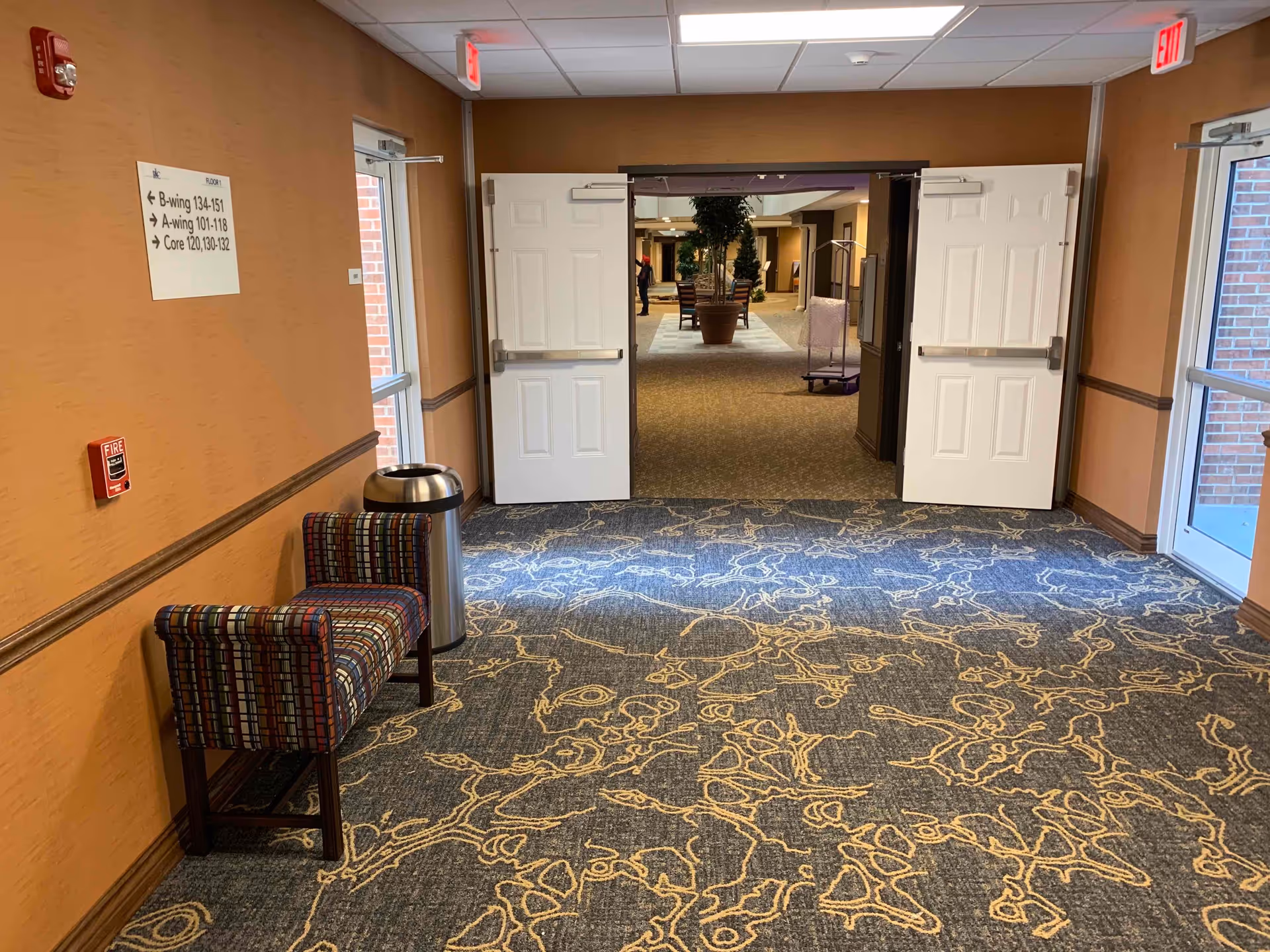 Interior hallway of Hudson Meadows Retirement Community with patterned carpet, a small upholstered bench, a trash can, and double doors leading to another hallway with plants and seating visible.