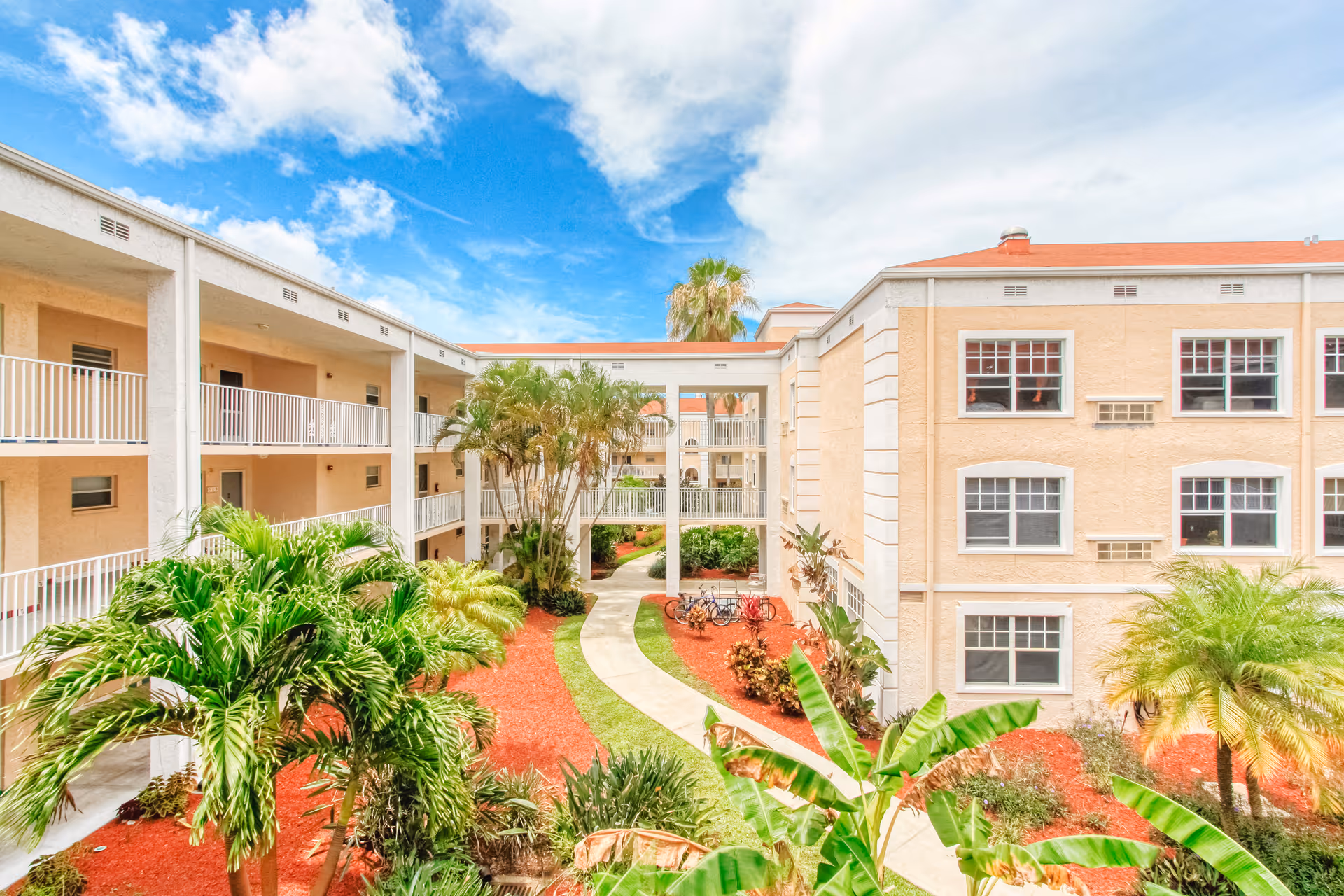 Outdoor courtyard area of a senior living facility with a winding concrete pathway, tropical plants, and palm trees surrounded by a three-story beige building with white railings and multiple windows under a partly cloudy blue sky.