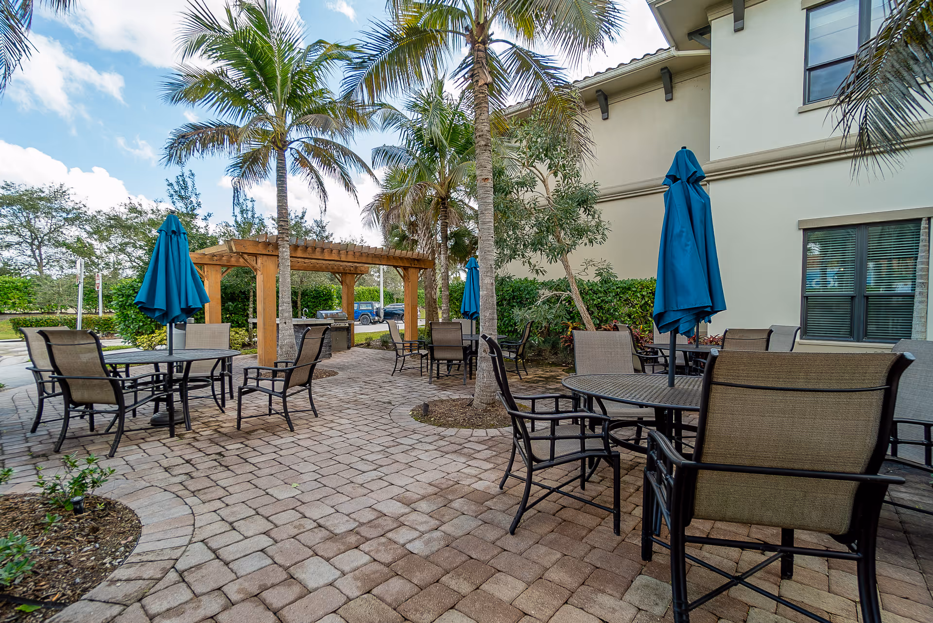 Outdoor patio area with several round tables and chairs, each table having a closed blue umbrella. The patio is paved with stone tiles and surrounded by palm trees and greenery. A wooden pergola is visible in the background near the edge of the patio, adjacent to a building with windows.