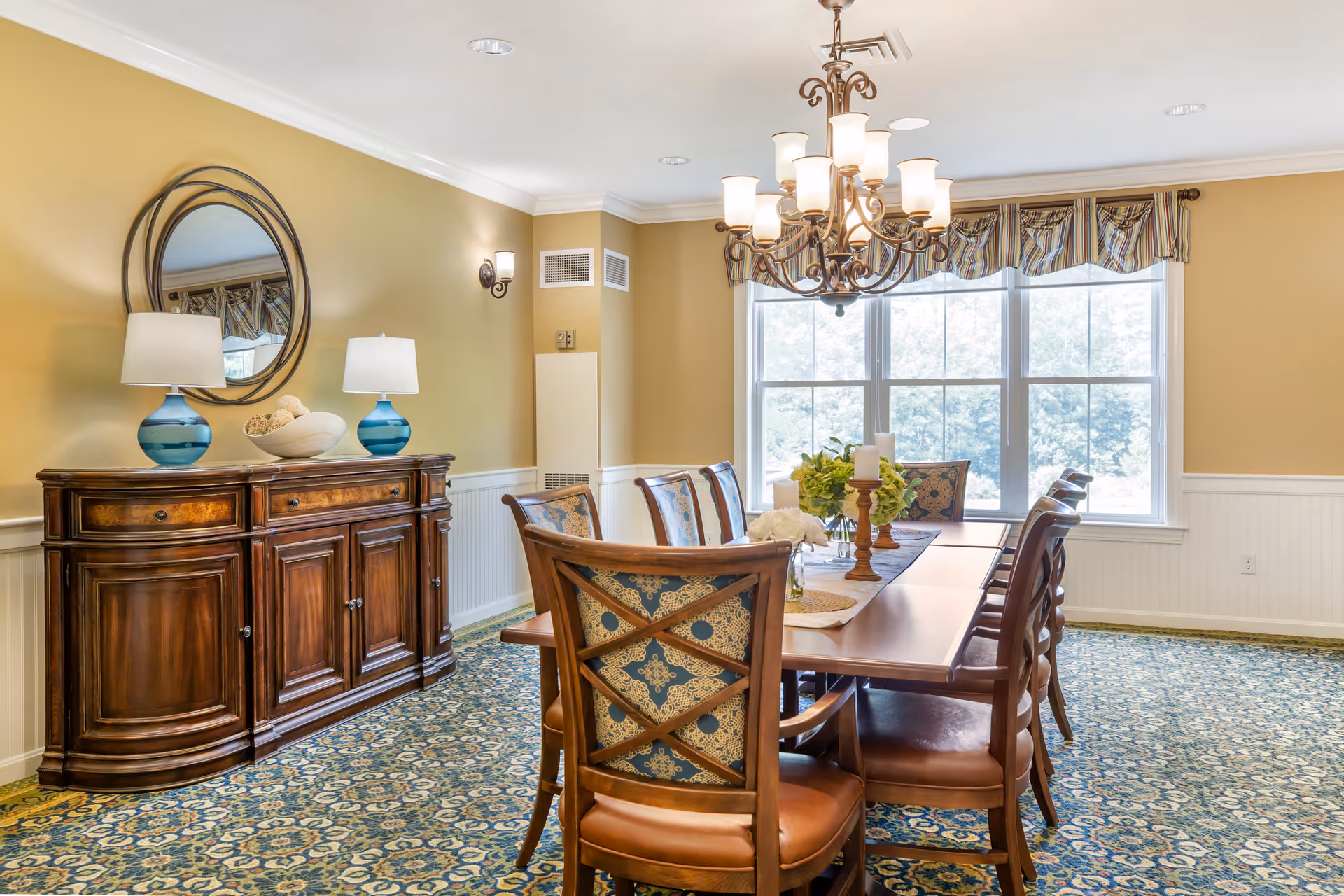 A well-lit dining room with a large wooden dining table surrounded by eight chairs with patterned upholstery. A decorative chandelier hangs above the table. On the table are candles and floral arrangements. Against one wall is a wooden sideboard with two blue lamps and a decorative bowl. A round mirror hangs above the sideboard. The room has beige walls, a patterned carpet, and large windows with striped valances letting in natural light.