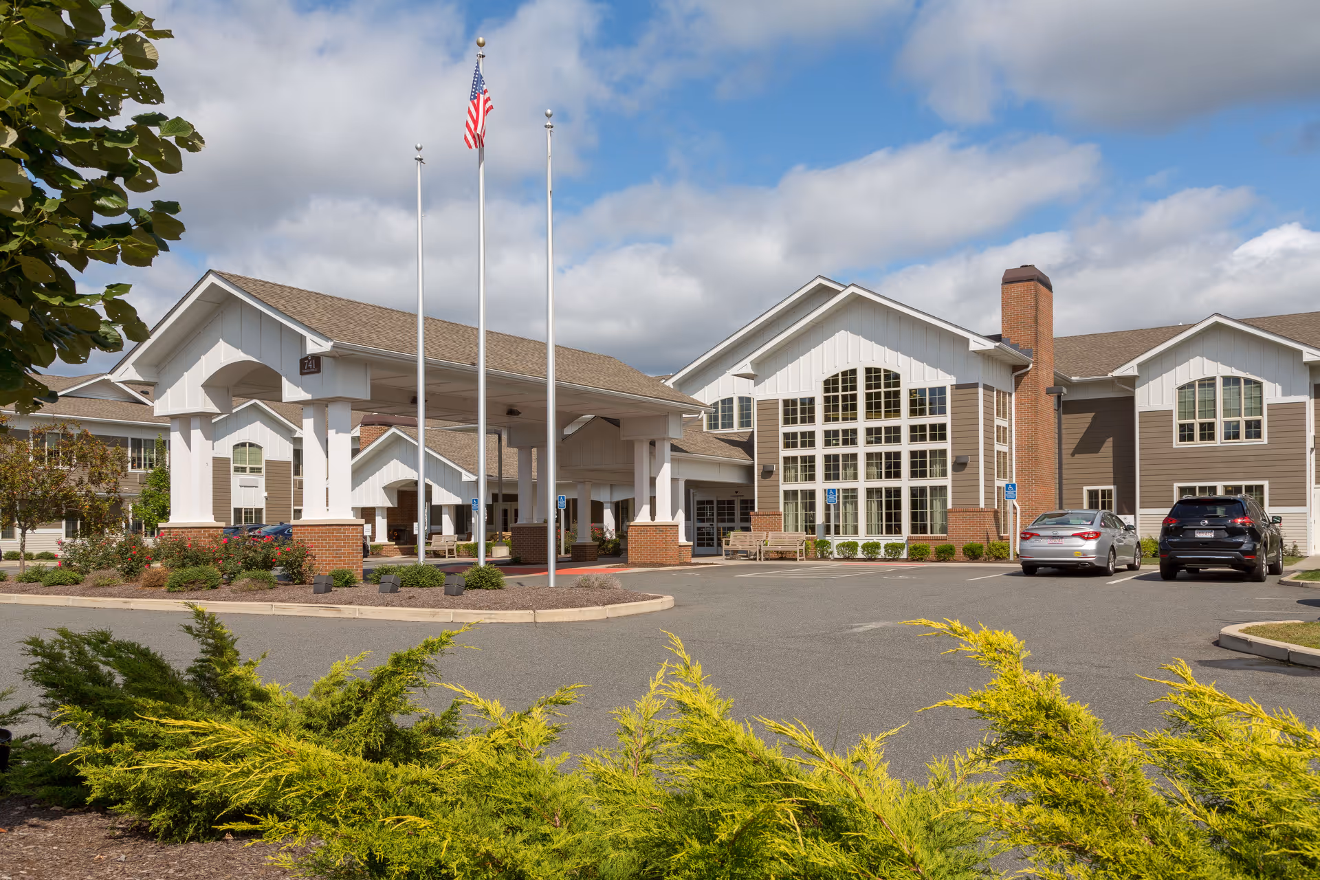 Front entrance of a senior living building with a covered porte-cochere, flagpoles, landscaped beds, and parked cars.