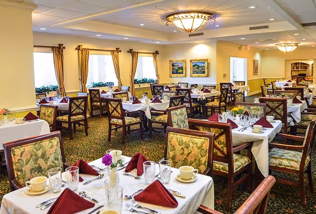 A bright dining room with multiple tables set for a meal, each covered with white tablecloths, burgundy folded napkins, cups, glasses, and silverware. The chairs have floral upholstery, and the room features large windows with beige curtains, framed landscape paintings on the walls, and warm ceiling lights.
