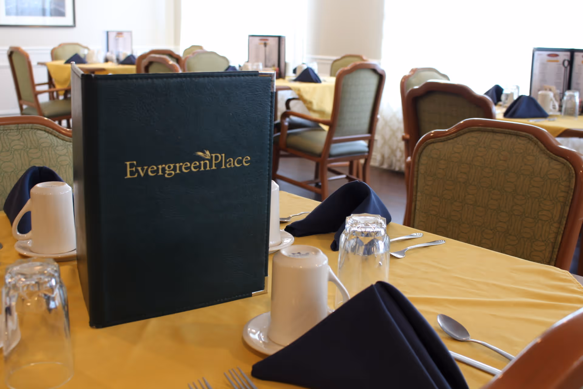 A dining table set with yellow tablecloth, navy blue folded napkins, white coffee mugs, upside-down drinking glasses, and silverware. A dark green menu with the text 'Evergreen Place' is prominently placed on the table. In the background, more tables and chairs are visible in a well-lit dining room.