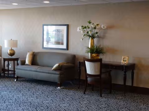 A furnished living room lobby with sofa, chair, desk, framed art, and flowers.