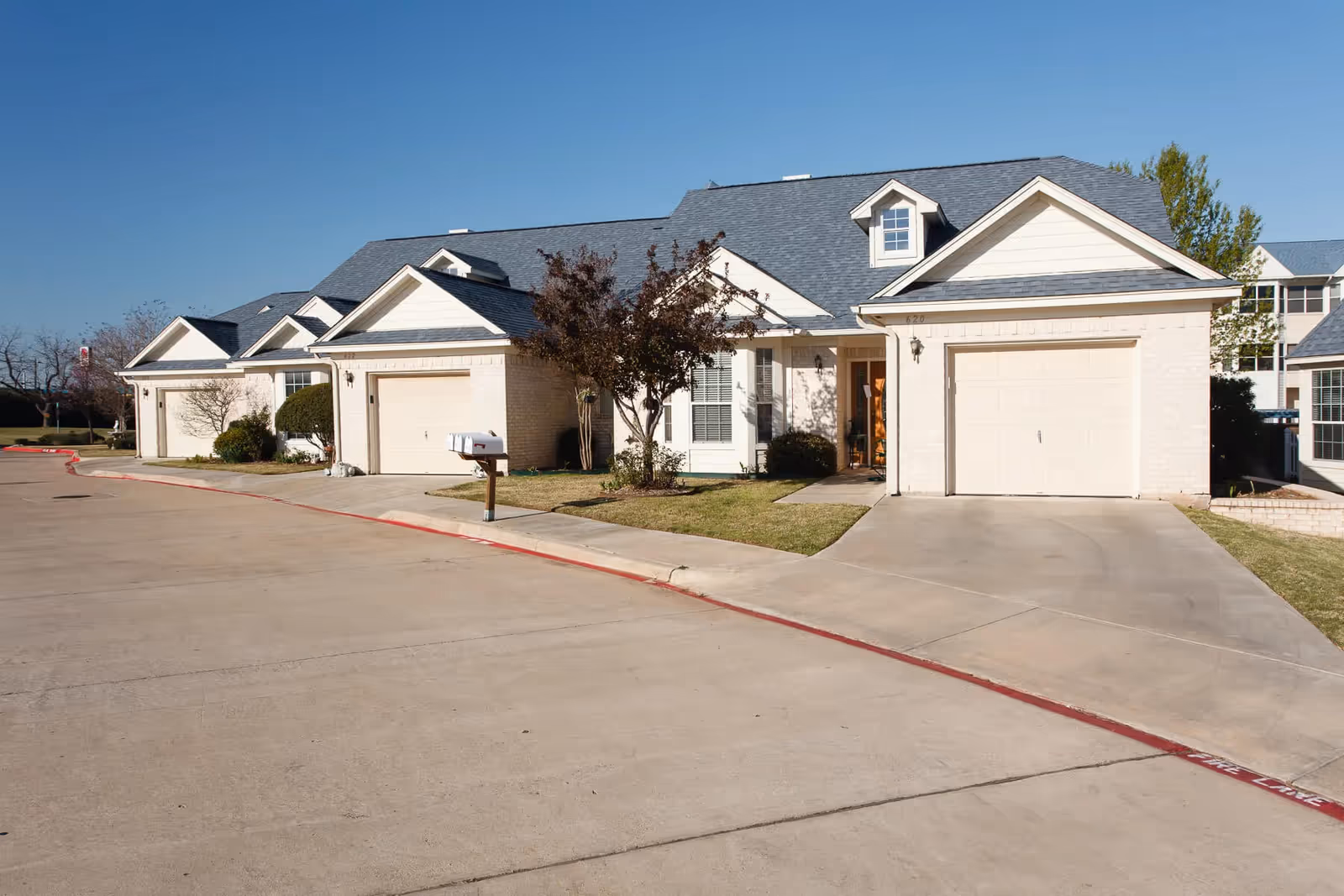 Exterior view of a single-story residential building with attached garages, a driveway, and a small lawn with trees and shrubs under a clear blue sky.