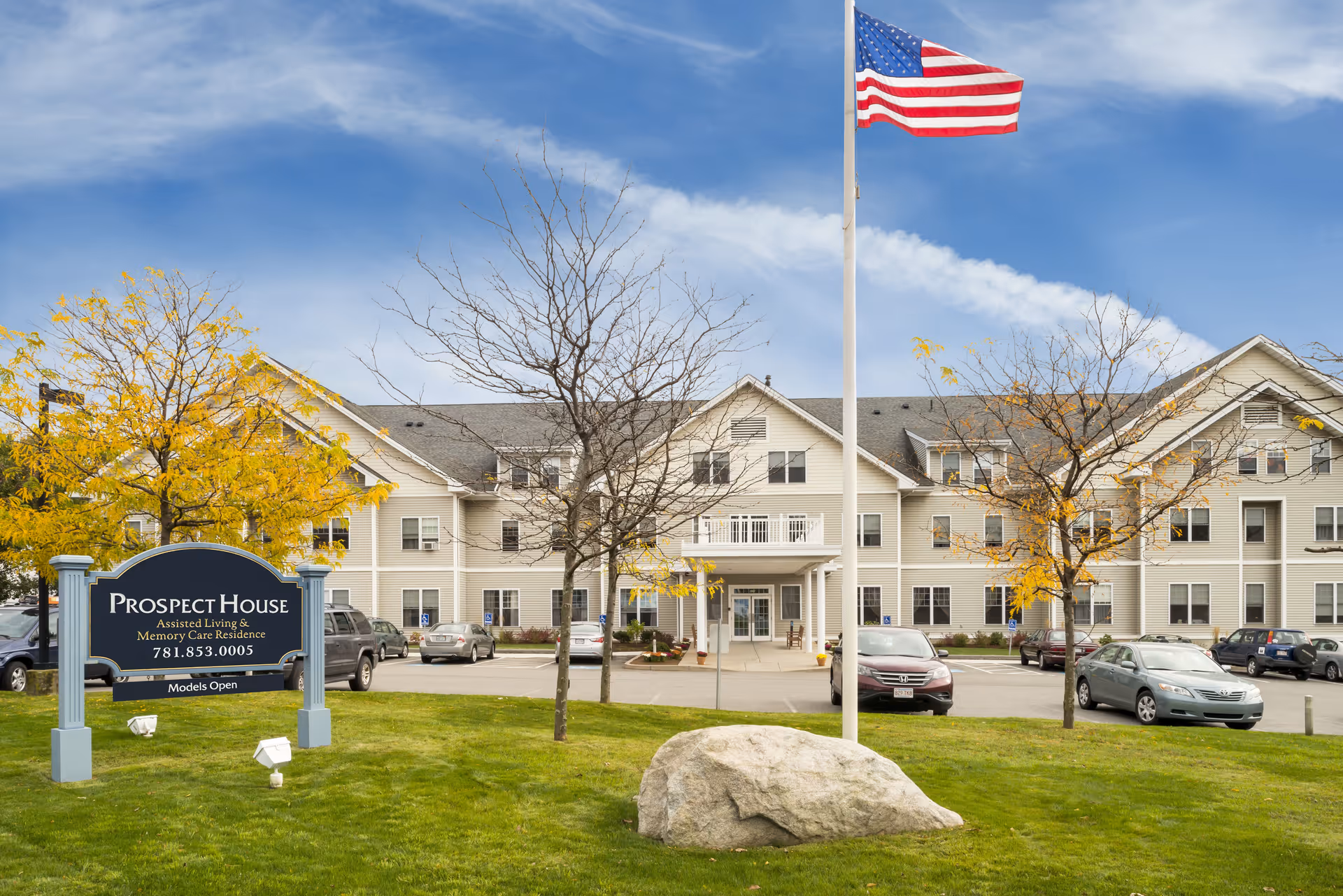 Exterior view of Prospect House Assisted Living and Memory Care Residence, a large beige multi-story building with several windows. In front of the building is a green lawn with a large rock, a flagpole flying the American flag, and a sign displaying the facility's name and contact number. Several cars are parked in the parking lot surrounding the building. Trees with yellow and bare branches are scattered around the lawn.