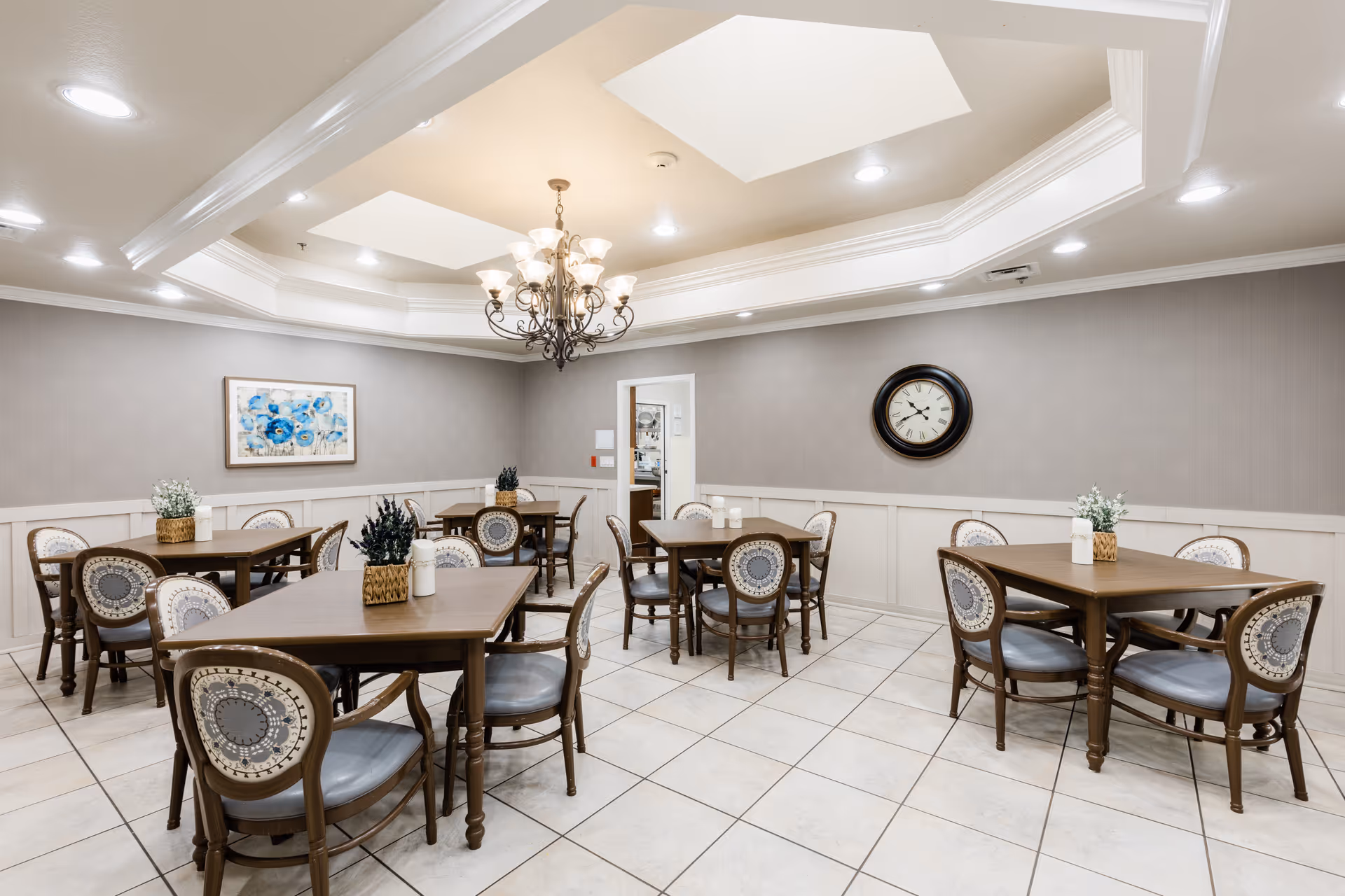 A dining room with several wooden tables and chairs arranged neatly. Each table has a small decorative plant. The room features a tiled floor, gray walls with white wainscoting, a large round clock on one wall, a framed floral painting on another, and a decorative chandelier hanging from a recessed ceiling.
