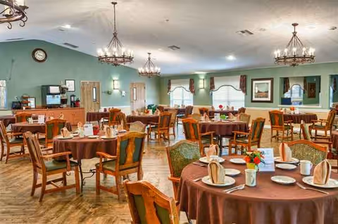 A spacious senior living dining hall with round tables, brown tablecloths, and chandeliers.