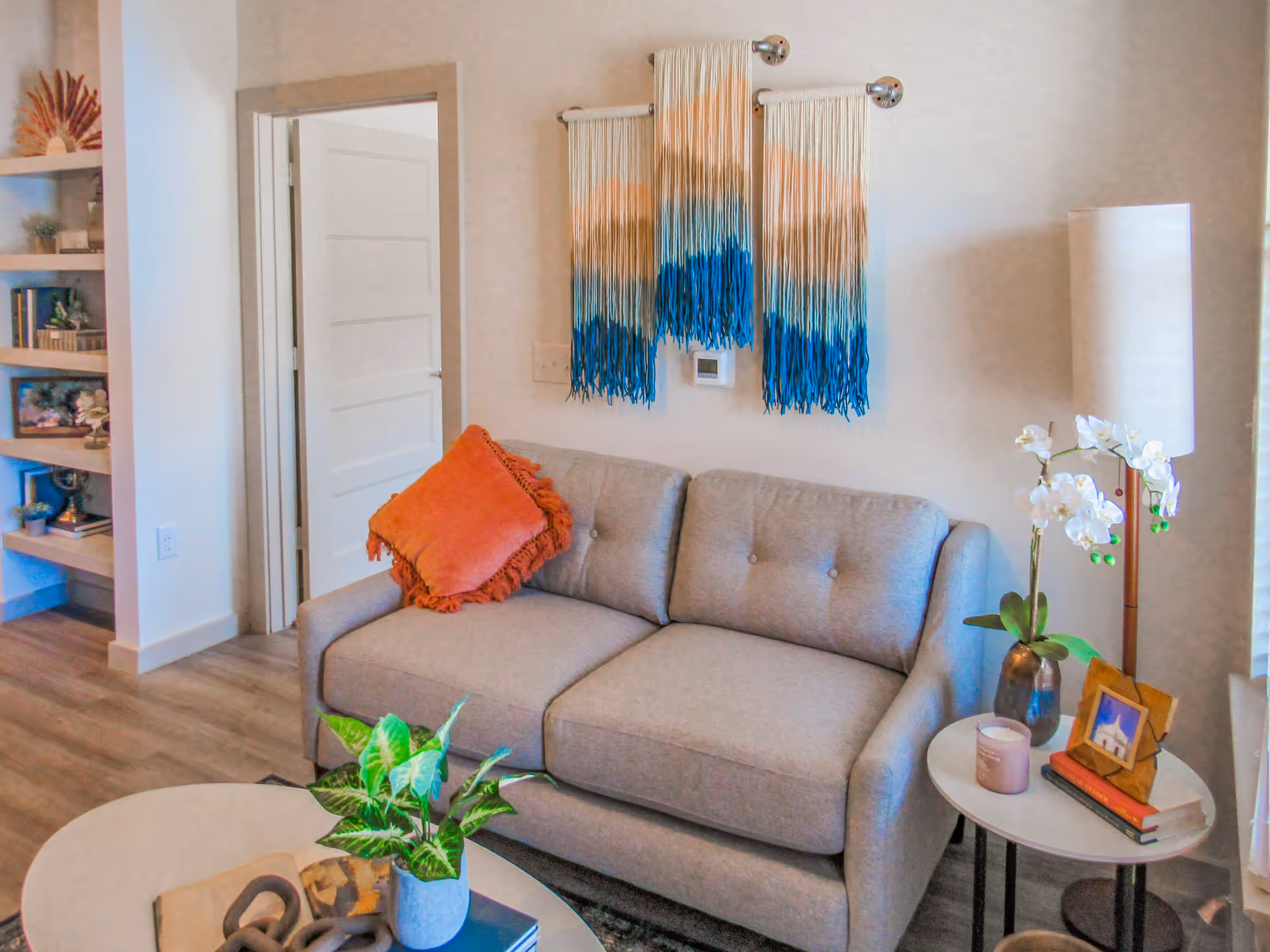 Small living room with a gray loveseat, woven wall hanging, coffee table with a plant, and a side table with a lamp and orchid.