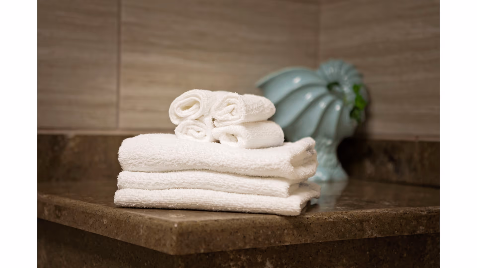 A stack of neatly folded white towels placed on a brown countertop with a decorative light blue ceramic shell-shaped vase in the background.