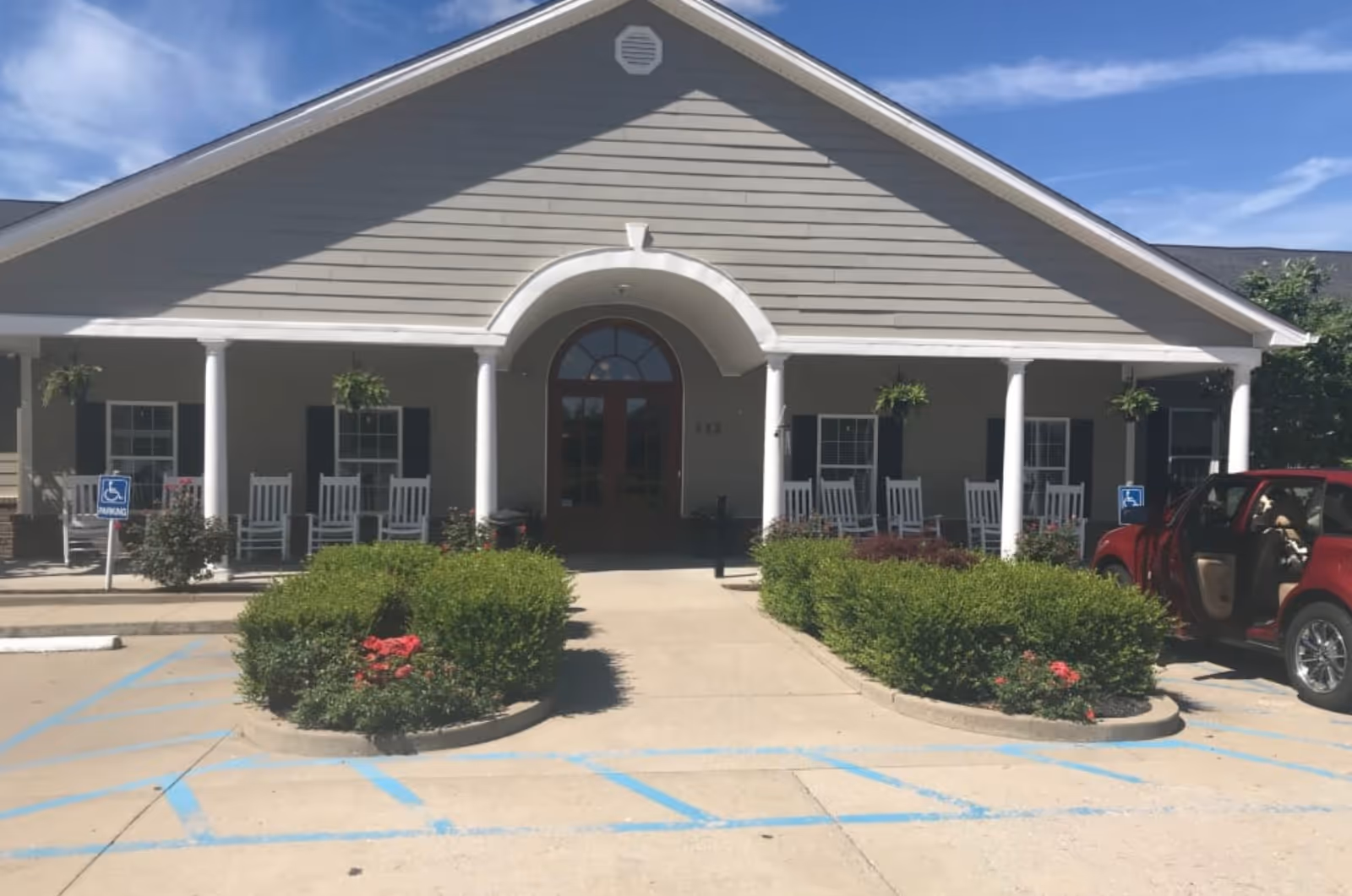 Front exterior view of a single-story building with a covered porch featuring white rocking chairs and hanging plants. There are two handicap parking spaces with blue markings and signs in front of the building. A red car is parked on the right side with its door open. The sky is clear with some clouds.