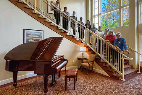 A group of people standing on a staircase inside a building with large windows letting in natural light. In the foreground, there is a grand piano with a bench, a small table with a lamp, and an armchair. The carpet has a patterned design.