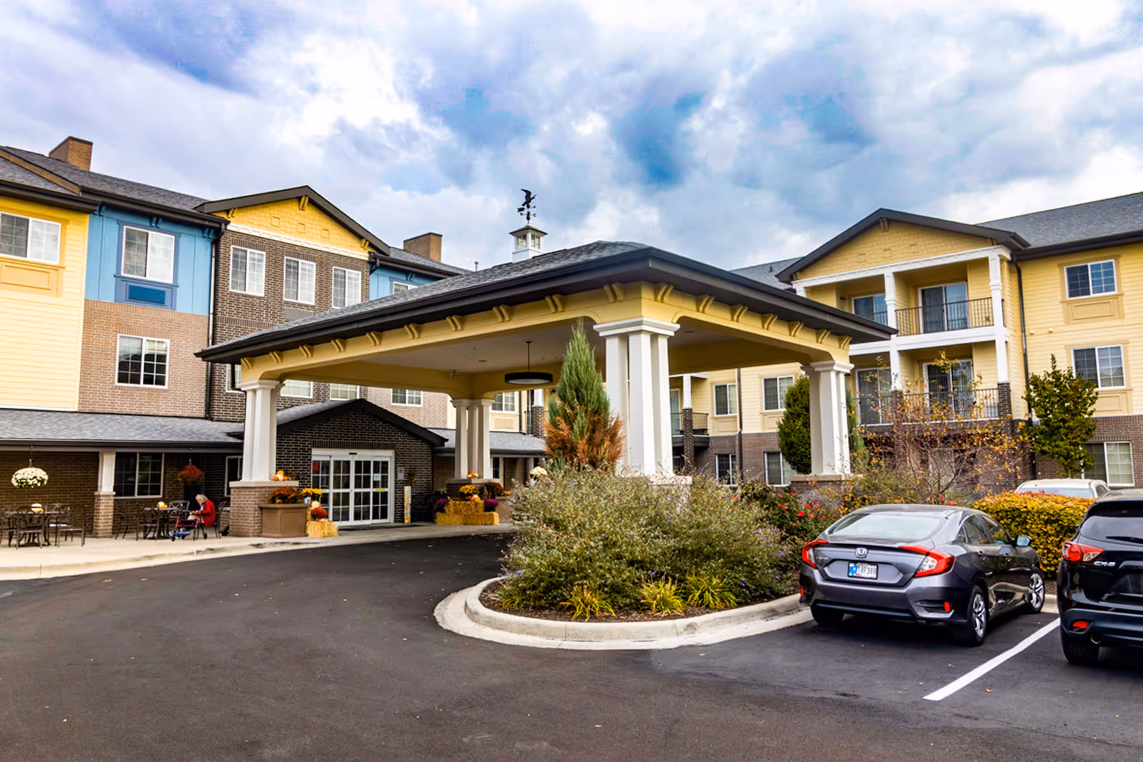 Covered porte-cochère entrance of a multi-story senior living building with parked cars and landscaping.