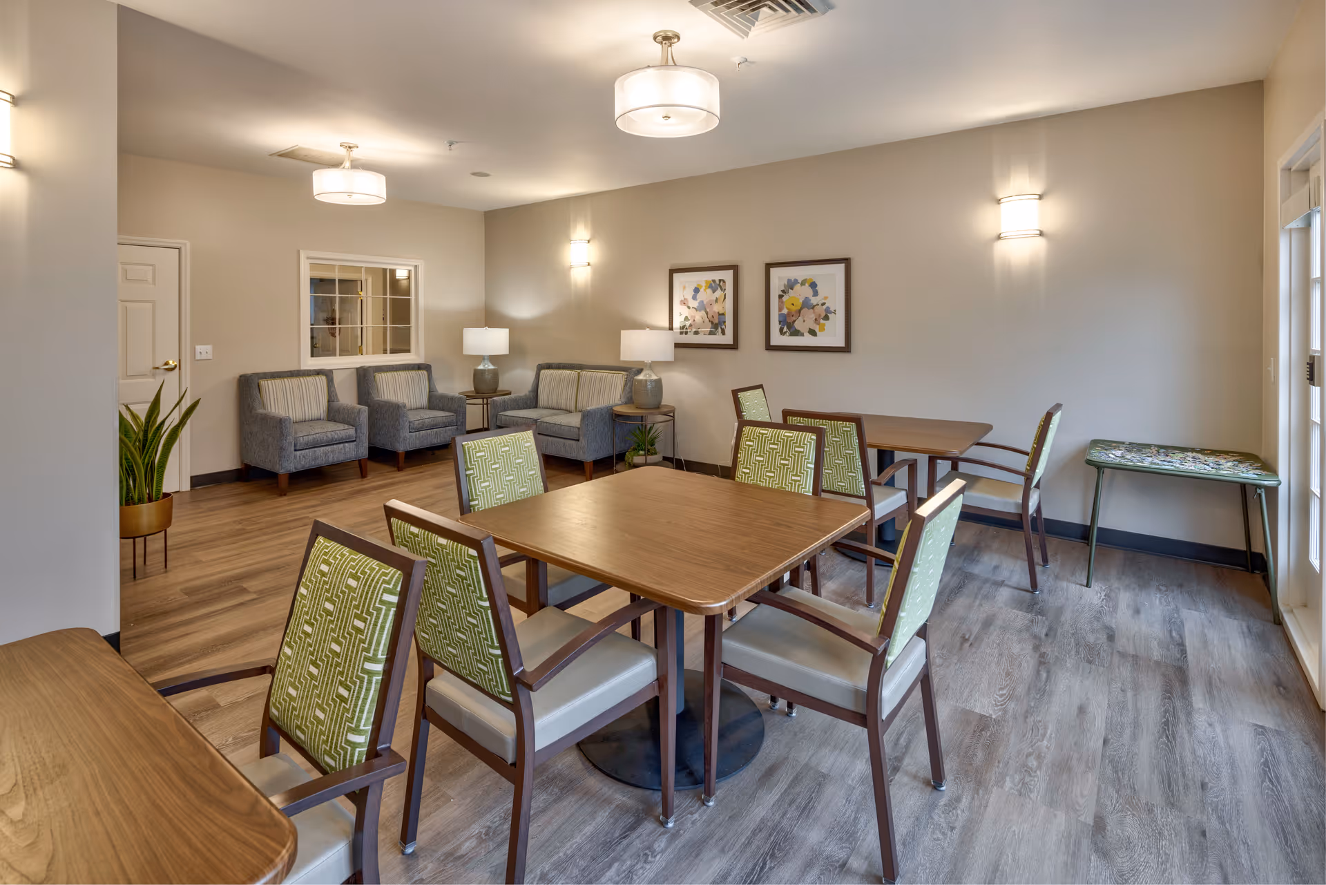 A well-lit room with wooden tables and chairs featuring green patterned upholstery. In the background, there are three gray armchairs with striped cushions, two table lamps on side tables, framed floral artwork on the wall, and a small plant in a pot. The room has wood flooring and neutral-colored walls with modern ceiling and wall light fixtures.