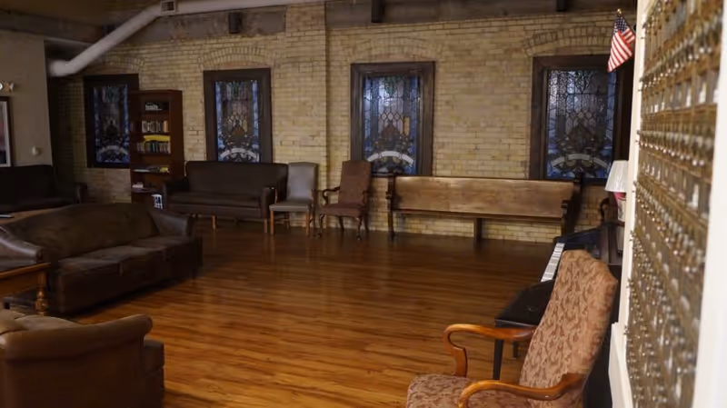 A spacious room with wooden flooring and exposed brick walls featuring three stained glass windows. The room is furnished with various seating options including leather sofas, wooden chairs, and a wooden bench. There is a bookshelf with books and an American flag in the corner. The right side of the image shows a wall with multiple mailboxes.