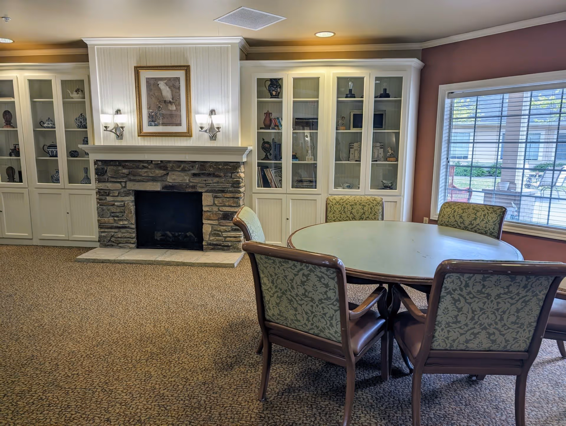 A cozy room with a round table surrounded by four upholstered chairs with patterned fabric. Behind the table is a stone fireplace with a white wooden mantel, flanked by built-in white cabinets with glass doors displaying decorative items. A large window with blinds lets in natural light, showing an outdoor view with patio furniture.