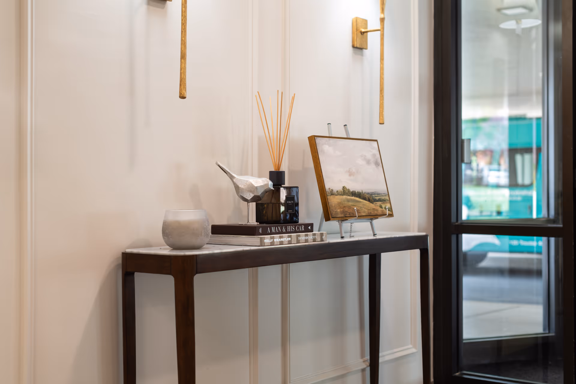 A narrow wooden console table with a marble top against a beige wall, decorated with a small silver bird figurine, a candle, reed diffuser, two stacked books, and a framed landscape painting on a stand. To the right is a glass door showing a blurred outdoor scene.