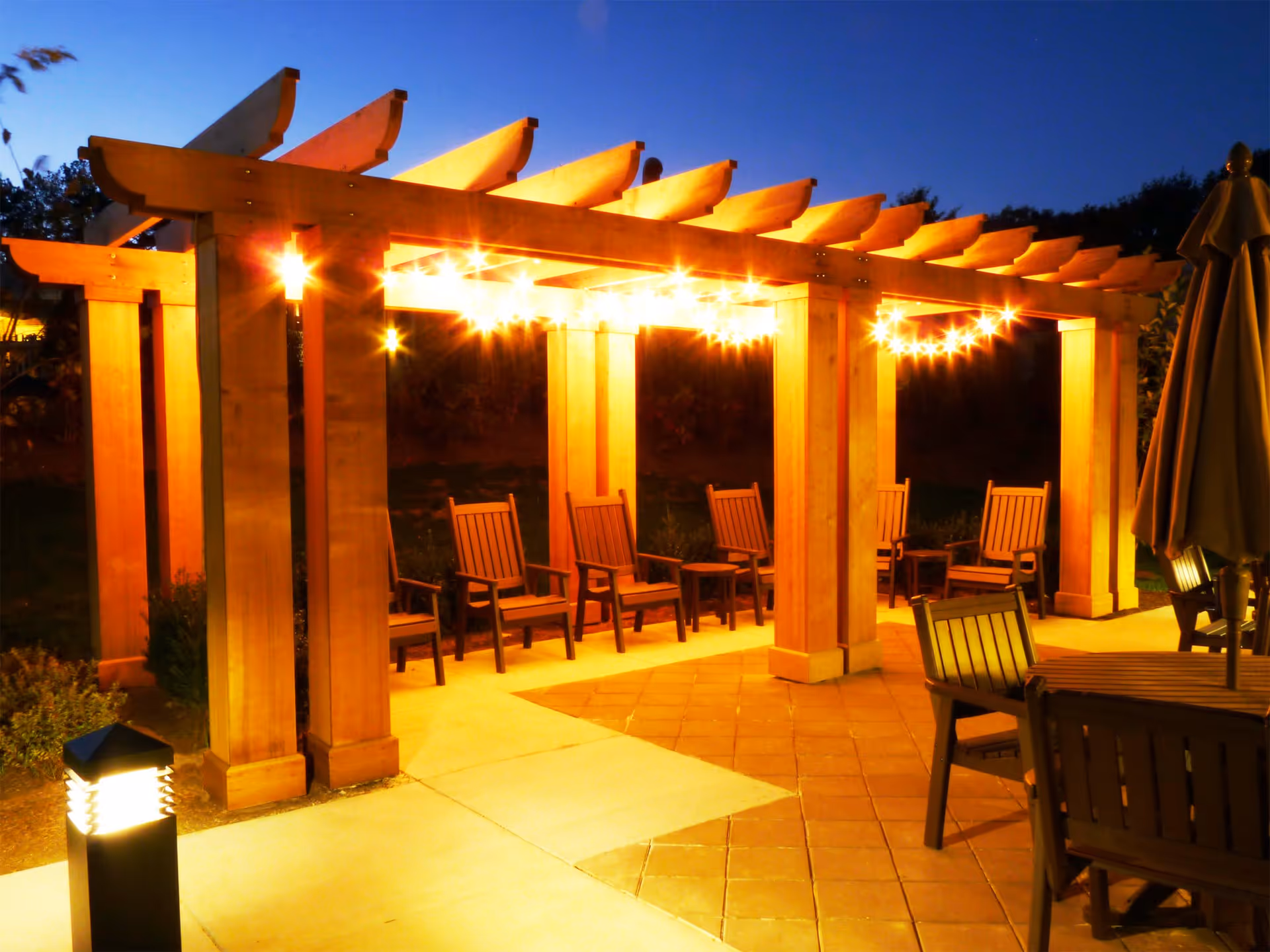 Outdoor seating area at dusk with a wooden pergola adorned with string lights. Several wooden chairs are arranged under the pergola and around a table with an umbrella. A lit pathway light is visible in the foreground.