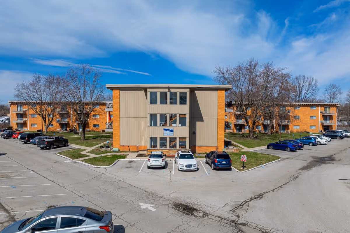 Exterior view of Pinnacle West Apartments, a three-story brick and beige panel building with several parked cars in front and leafless trees around the property under a partly cloudy blue sky.
