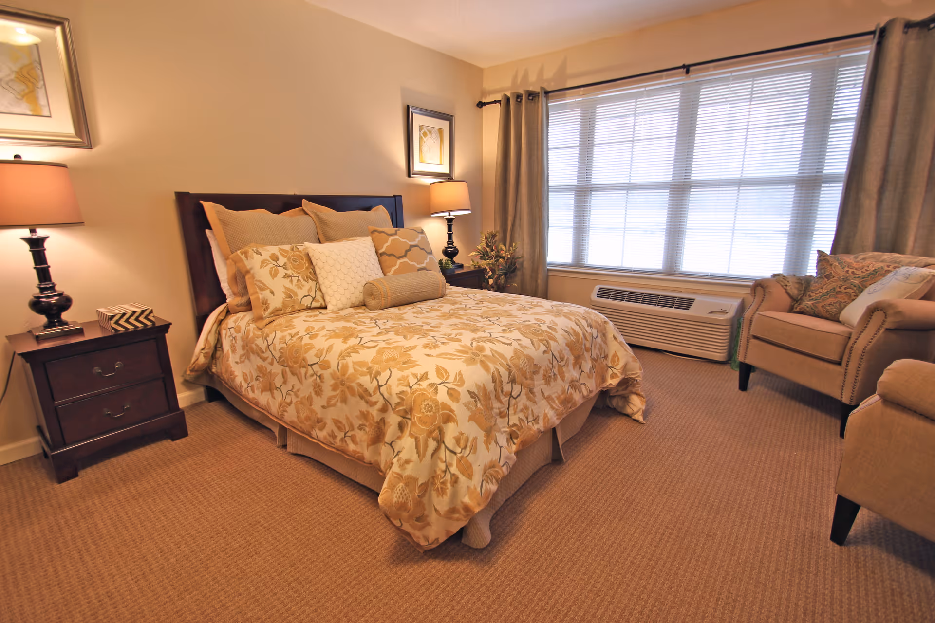 A cozy bedroom with a bed covered in a floral patterned comforter and multiple pillows. There are two bedside tables with lamps on each side of the bed. A large window with blinds and curtains lets in natural light. An upholstered armchair with decorative pillows is placed near the window. The room has beige walls and carpeted flooring.