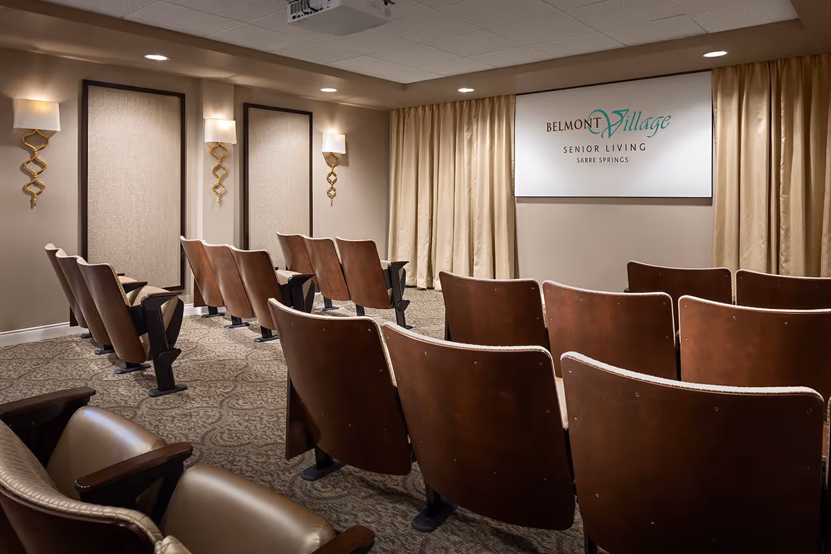 A small auditorium or presentation room with rows of vintage-style wooden theater seats facing a wall with a large sign that reads 'Belmont Village Senior Living Sabre Springs'. The room has beige walls, patterned carpet, three decorative wall sconces, and beige curtains behind the sign.