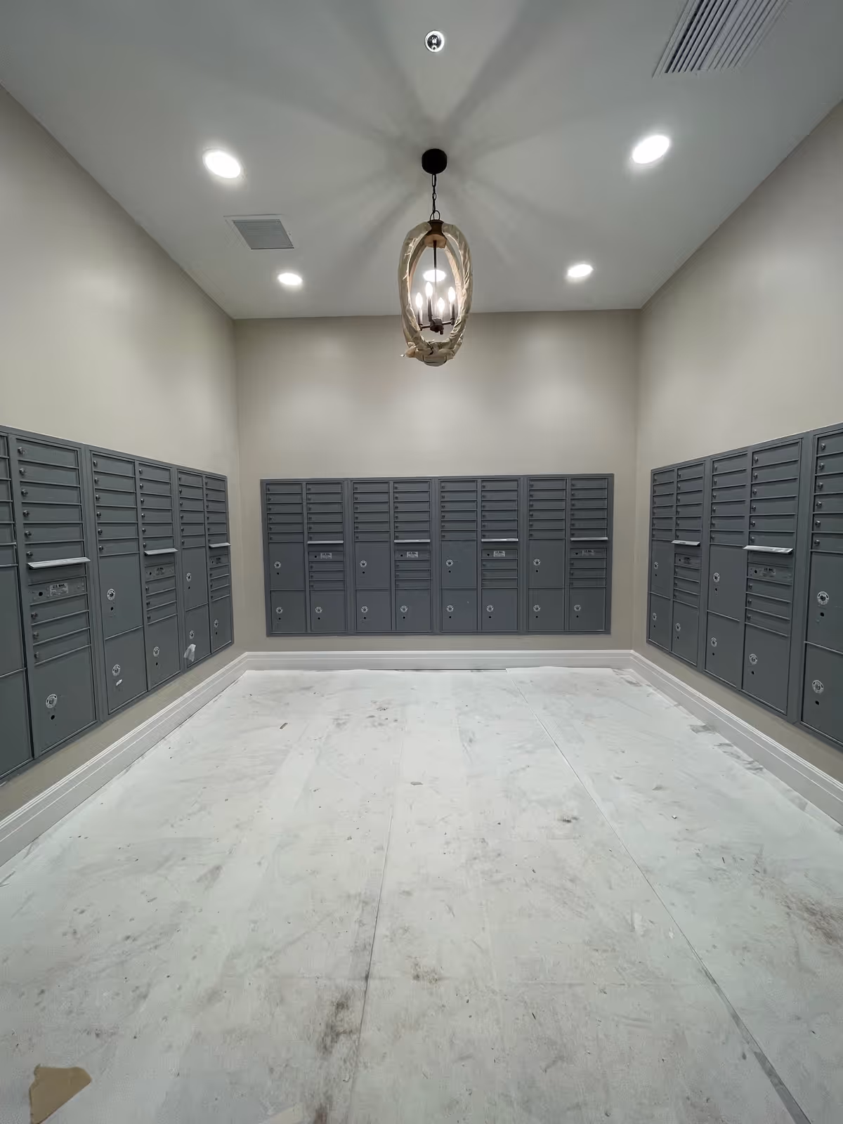 A room with multiple gray mailboxes mounted on three walls. The floor appears to be covered with a protective sheet, and a decorative ceiling light fixture hangs in the center of the ceiling. The walls are painted a light beige color, and there are recessed ceiling lights providing illumination.