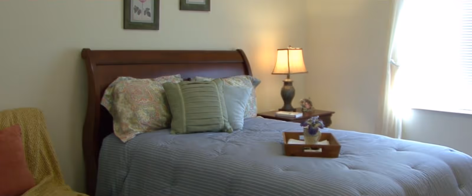 A cozy bedroom with a wooden sleigh bed made up with a blue striped comforter and several pillows in different patterns and colors. A wooden nightstand with a lamp and a small decorative item is beside the bed. A window with white curtains allows natural light to brighten the room. A chair with a yellow throw blanket and a red pillow is partially visible on the left side.