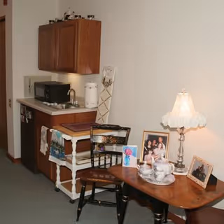 Small kitchenette and seating area with a sink, microwave, wooden cabinets, and a table holding family photos and a lamp.