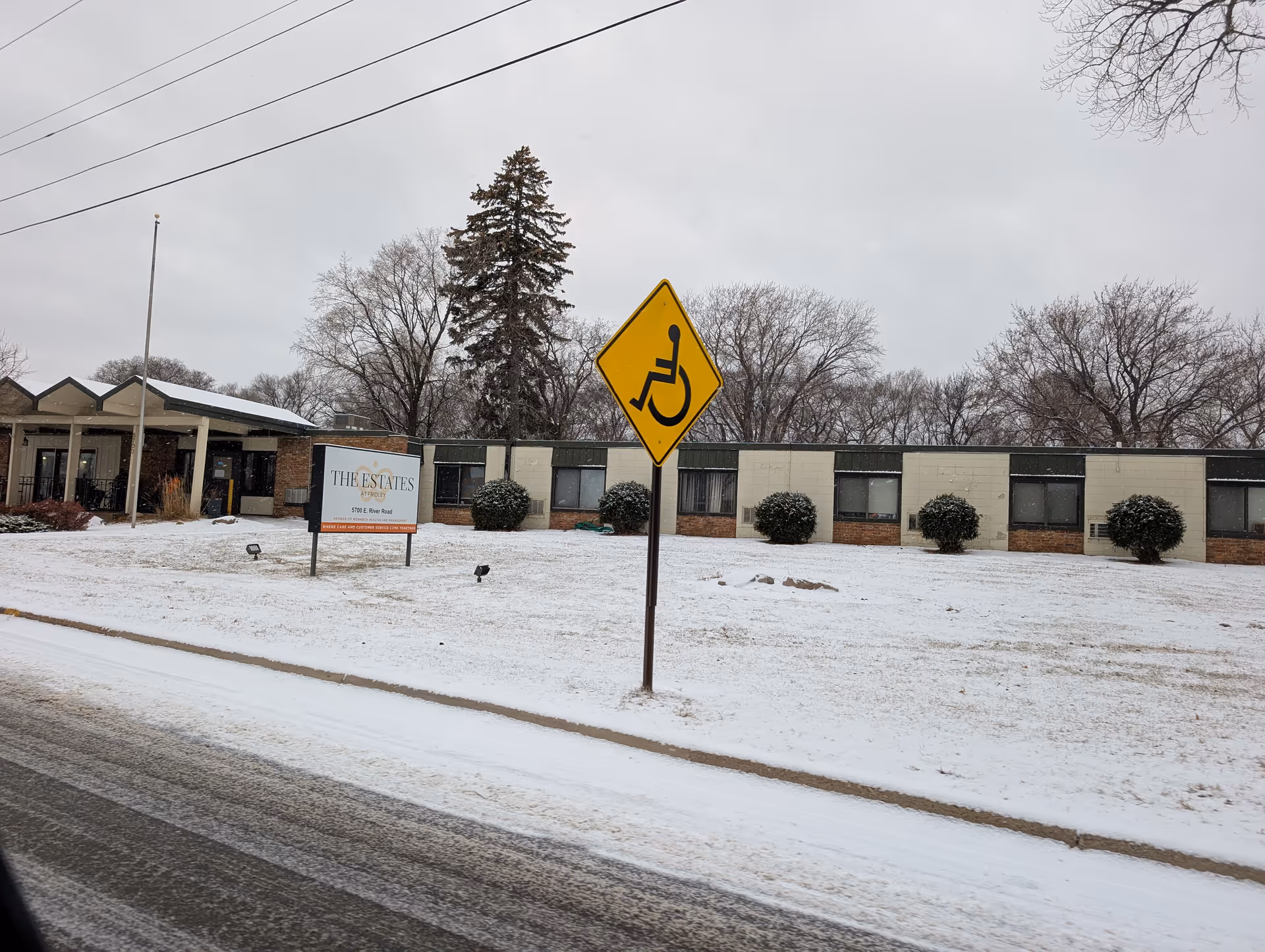 Front exterior of The Estates at Fridley building in winter with a handicap crossing sign and a snow-covered lawn.